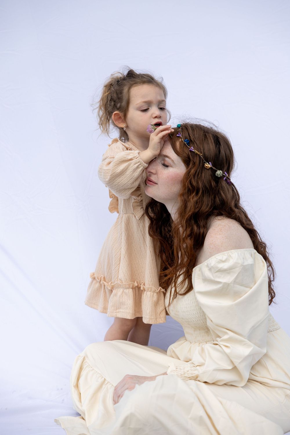 A woman in a cream-colored dress sits while a child in a beige ruffle dress stands beside her in a dreamy portrait setting.