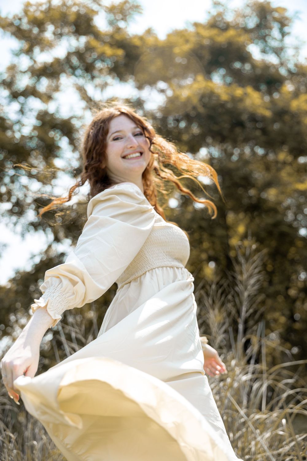 A joyful figure in a flowing white dress twirls outdoors with wind-swept hair in golden sunlight.