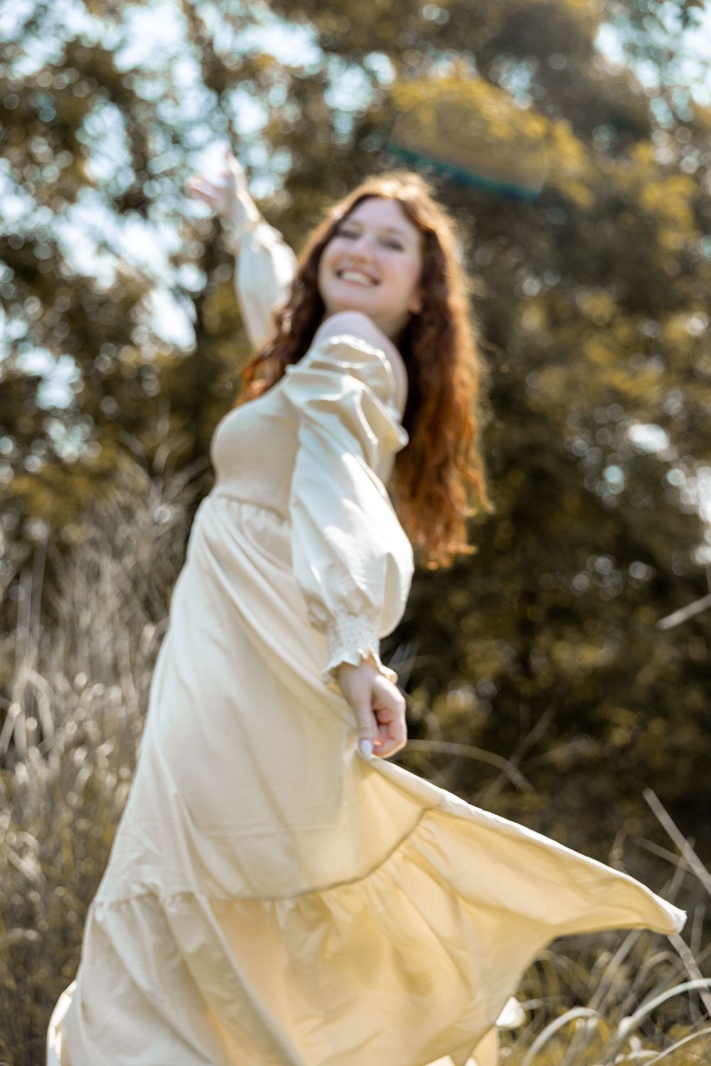 A person in a flowing cream dress twirls joyfully in a sun-dappled field of tall grass.