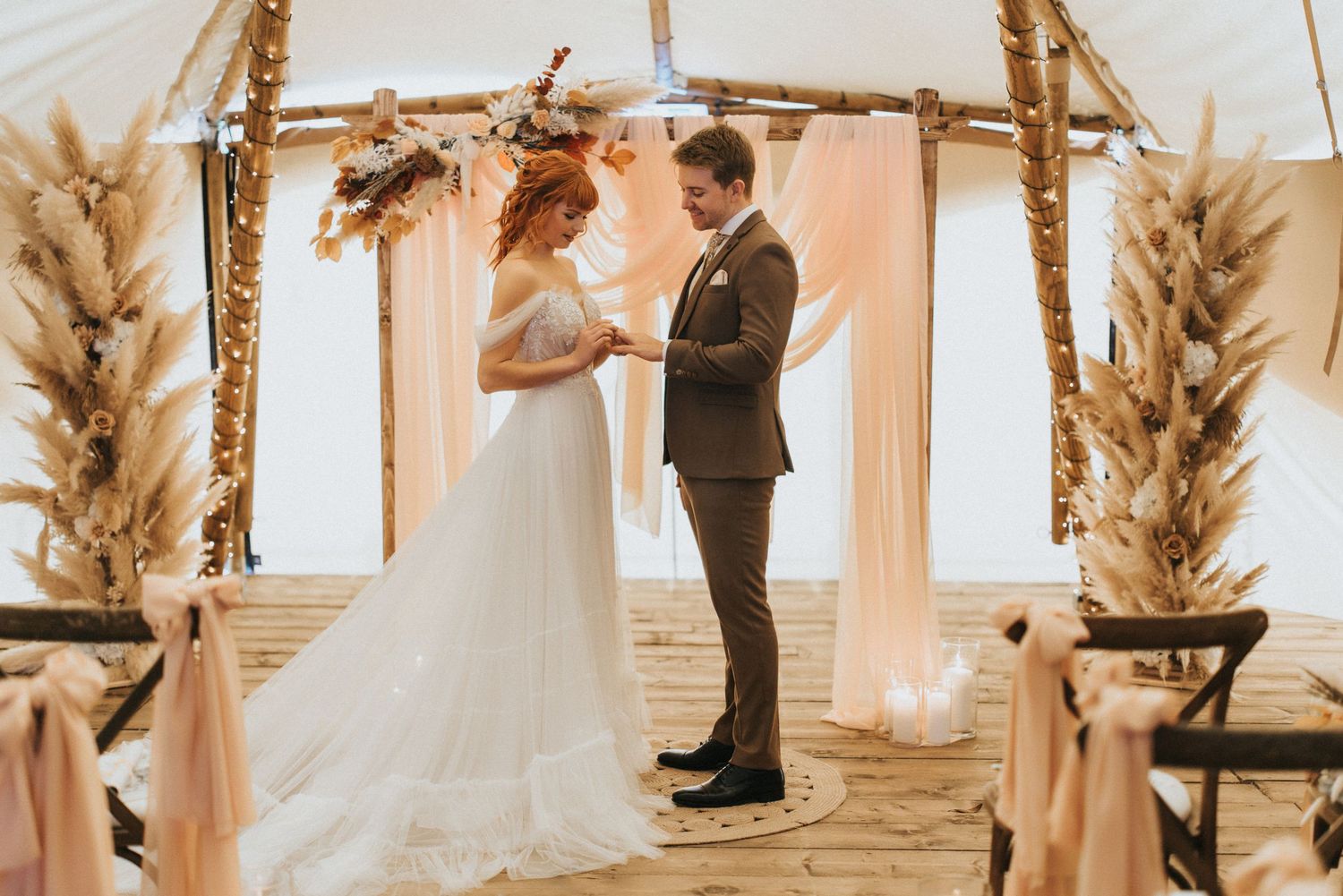 A wedding ceremony takes place under a rustic wooden arch decorated with pampas grass and soft peach drapery at Nuddock Wood Lake.
