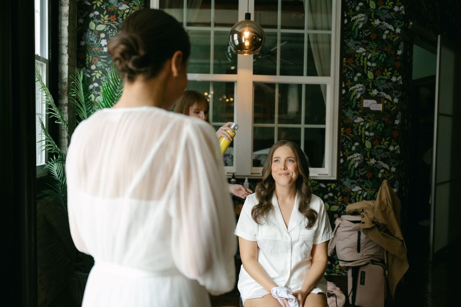 Two people sit and chat over coffee at a bright indoor cafe space with white windows and green plants.