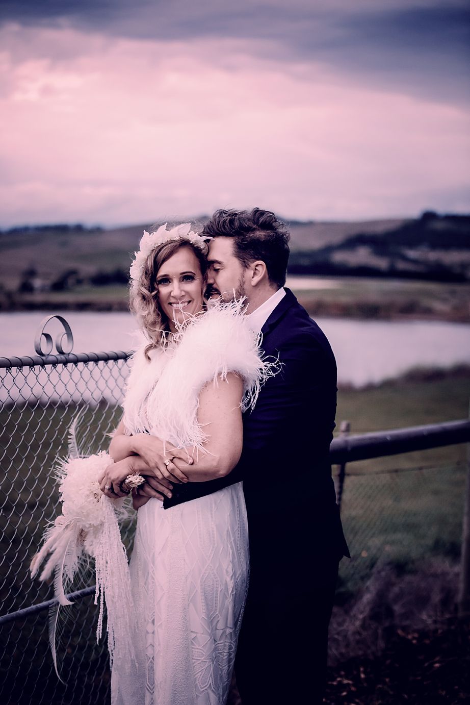 A romantic couple in formal attire embrace against a twilight landscape backdrop during a wedding photoshoot.