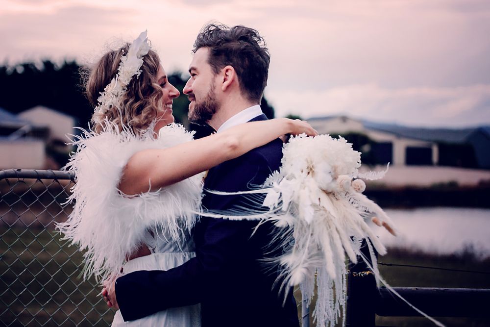 A couple in wedding attire wearing white feather wings embrace by a lake at sunset.