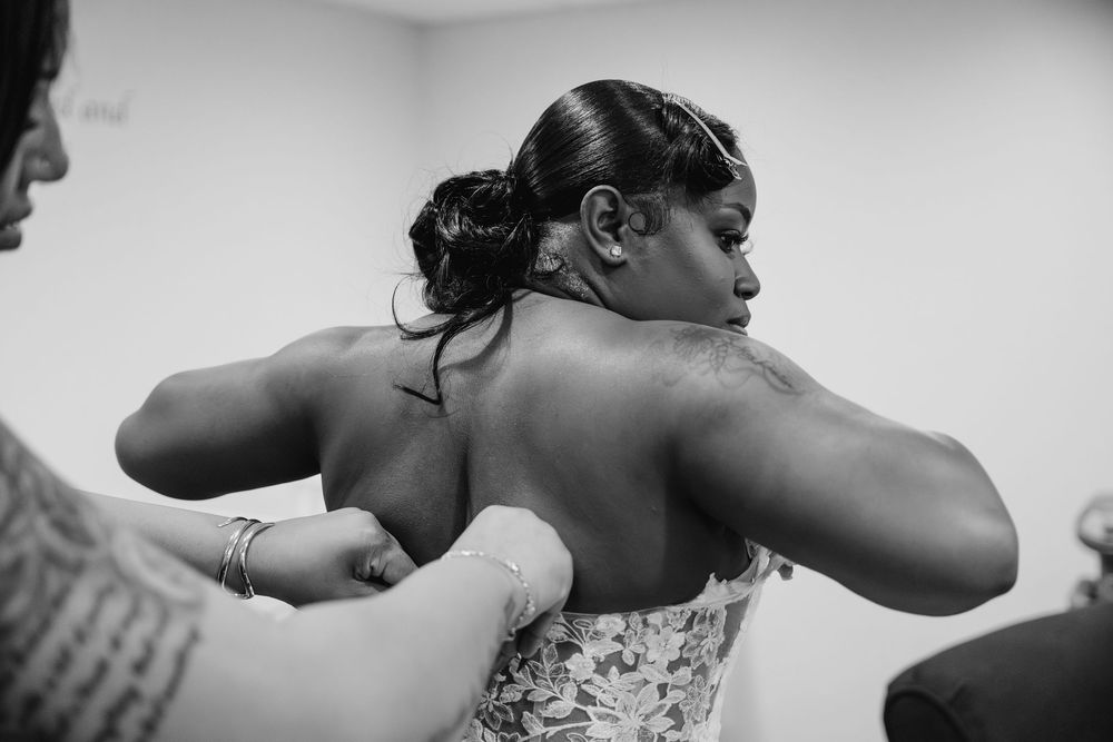 A black and white photo shows someone being helped into a wedding dress with lace details during preparation.