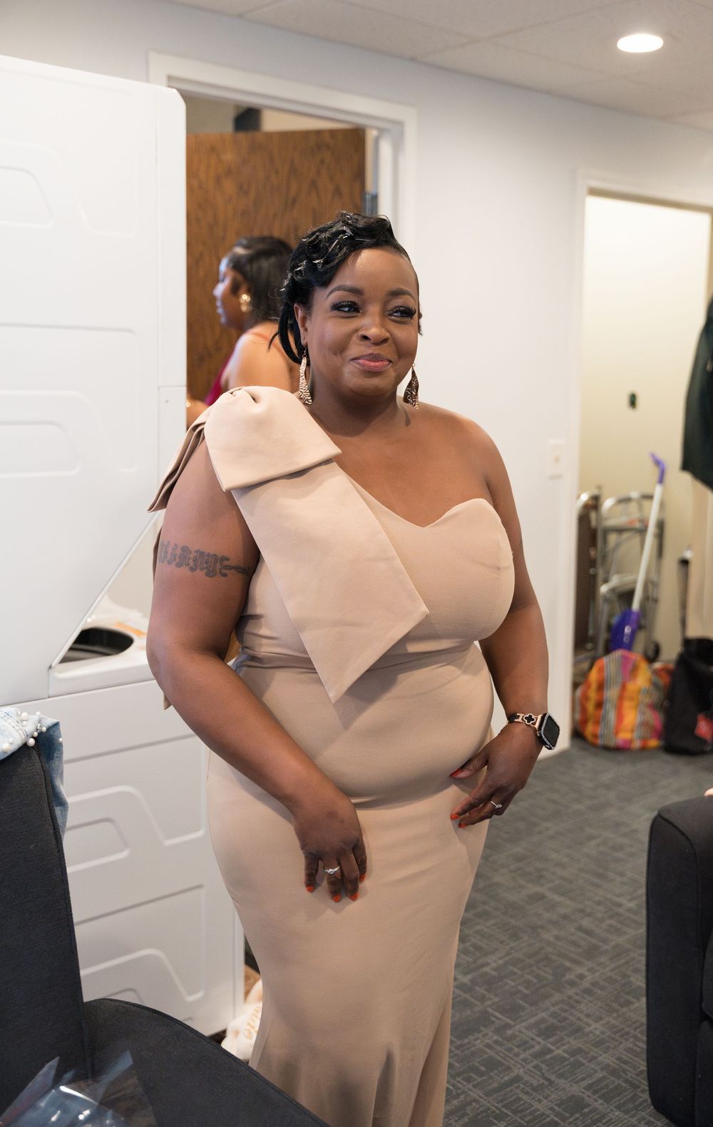 Woman in a light beige fitted dress poses against a white wall in an indoor setting.