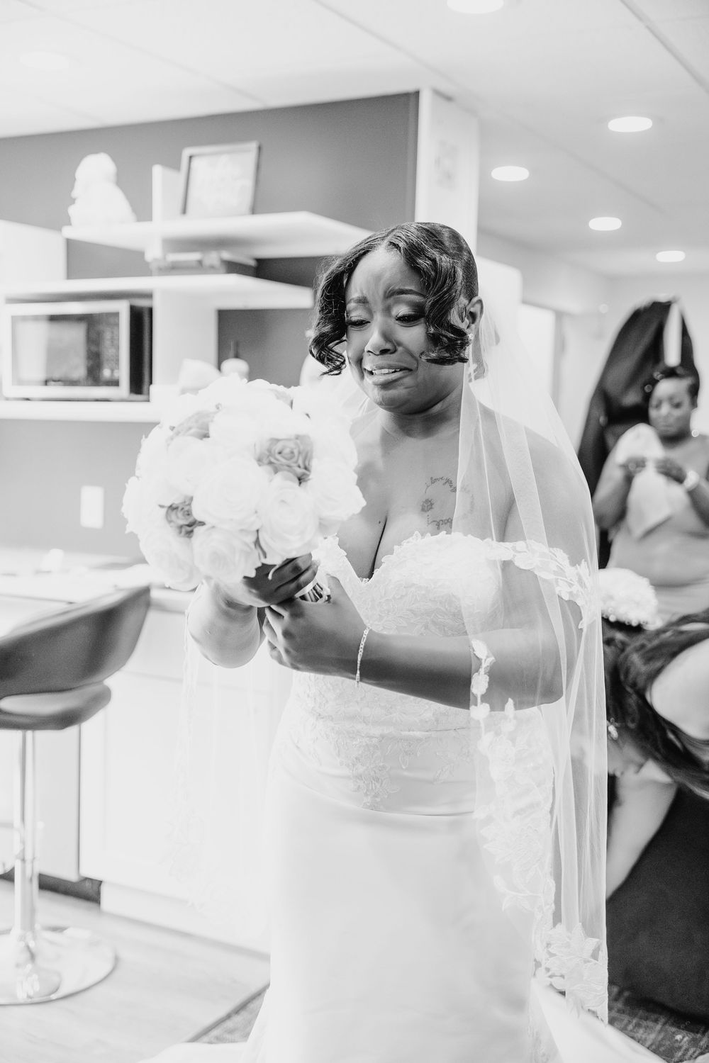 A bride in an elegant white wedding gown and veil holds a delicate bouquet during preparations in a bridal suite.