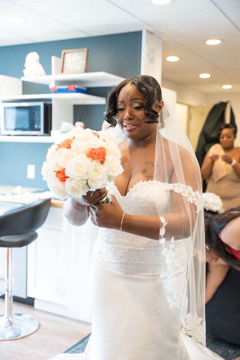 A beautiful bride in a white lace wedding dress and veil holds a peach and white bouquet while getting ready.