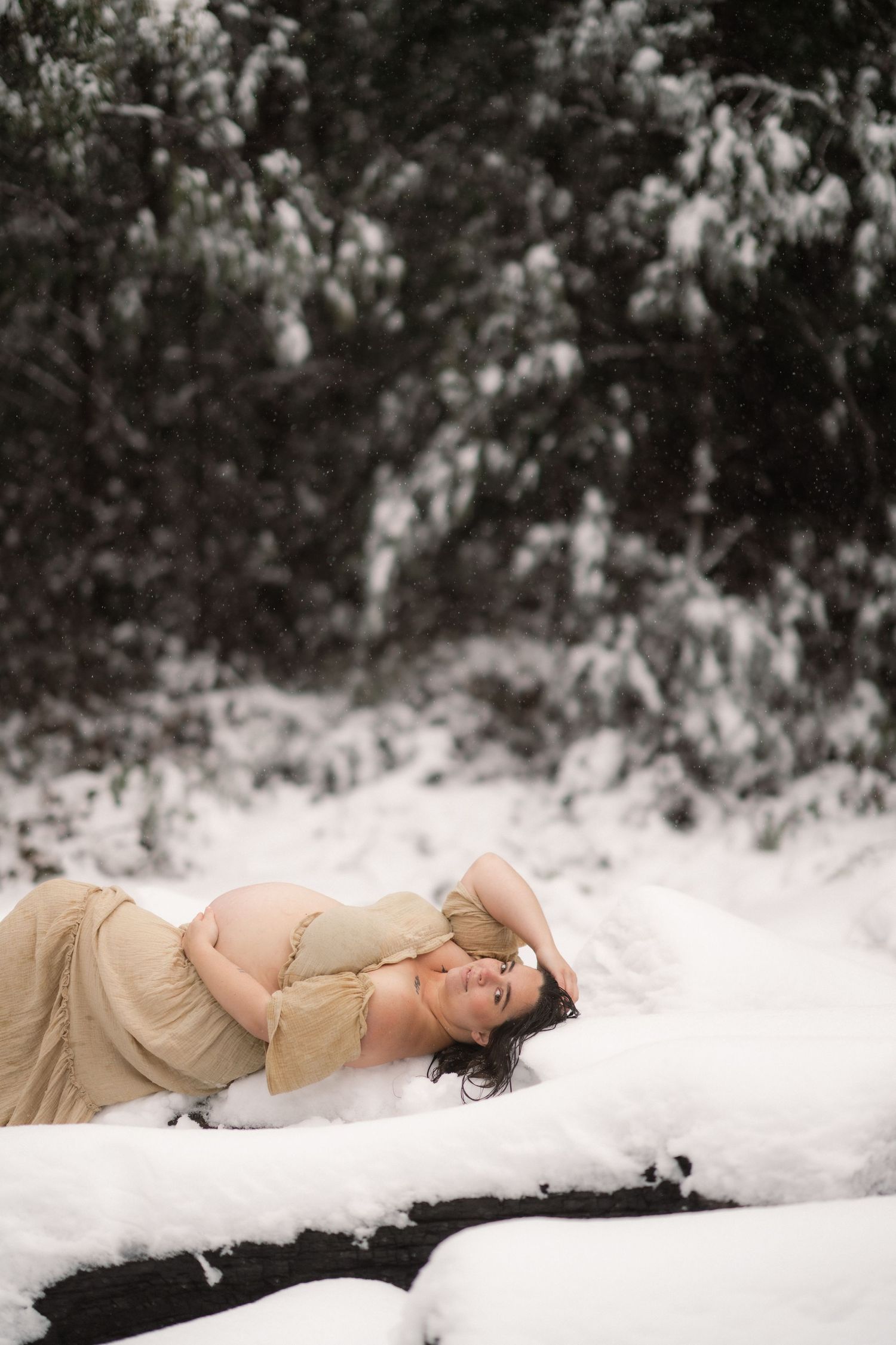 Ethereal winter scene with someone in flowing beige dress lying peacefully in pristine snow against evergreen backdrop.