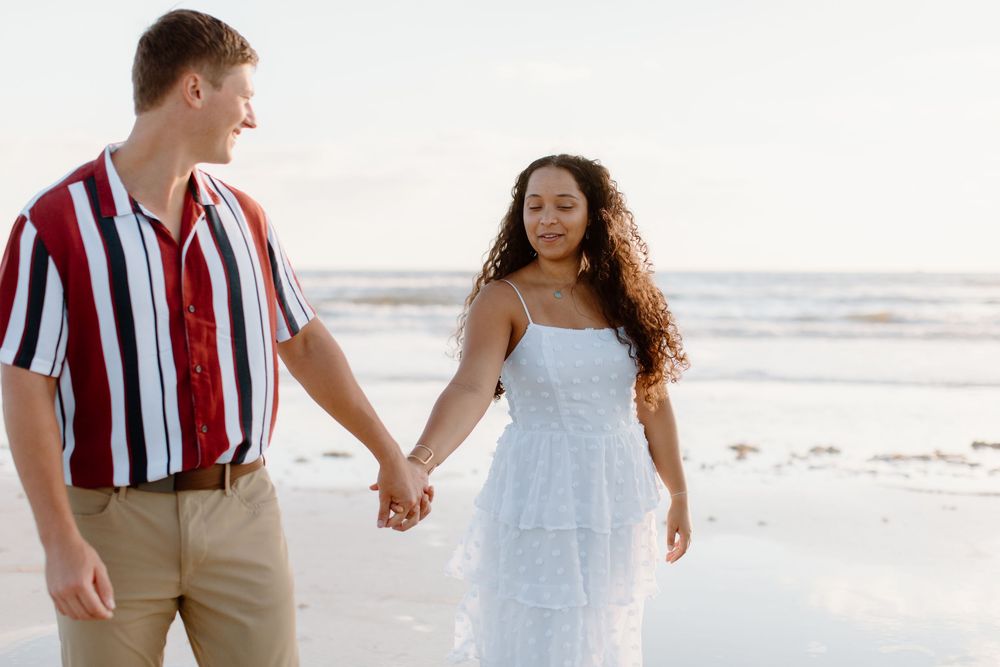 Couple walking hand in hand on beach, wearing white dress and striped shirt.