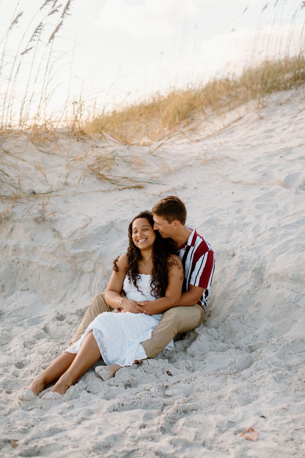 Couple sitting together on sand dunes during golden hour photoshoot.