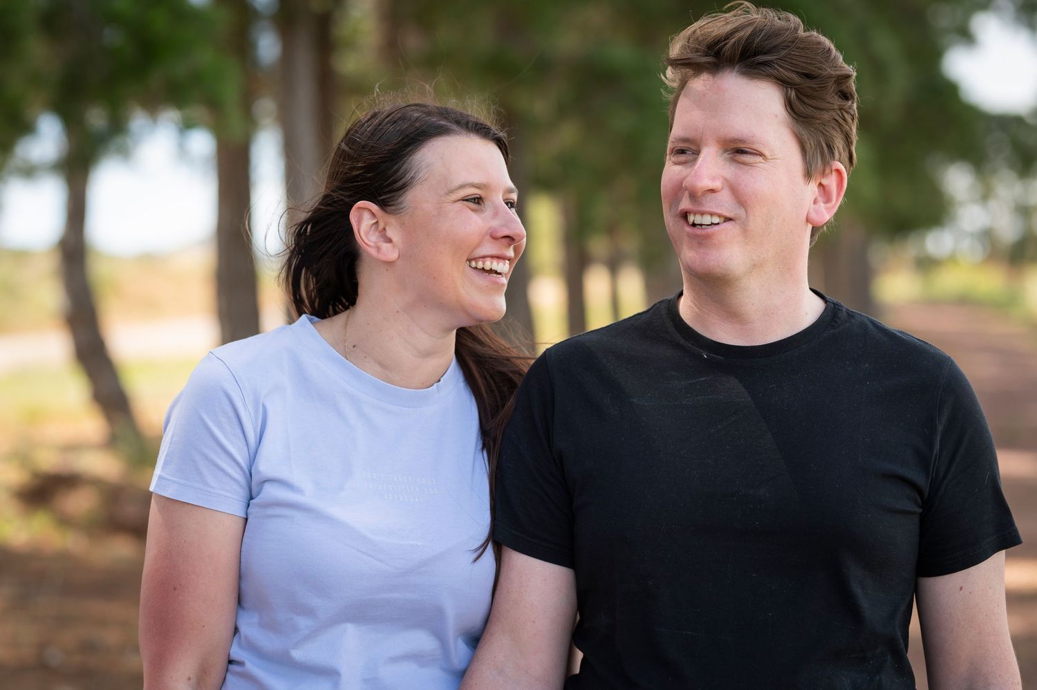 A young couple wearing t-shirts laughs together while walking outdoors in a sunny wooded area.