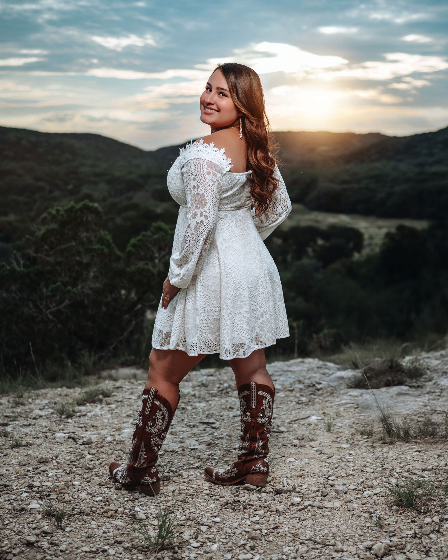 Person in red top and denim shorts sitting on rocky overlook at sunset with mountains in background.