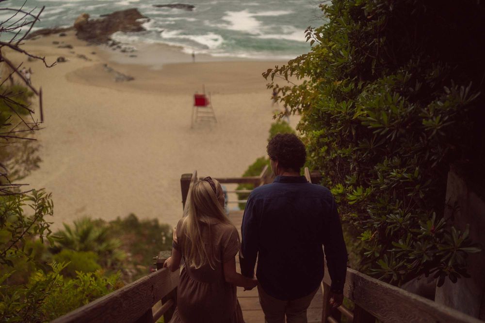 A couple walks down wooden stairs to a misty beach with a red umbrella visible on the sand in the distance.