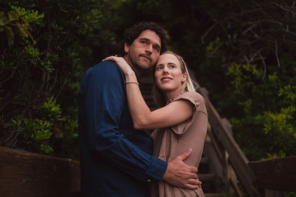 A couple embraces lovingly outdoors during a warm glowing sunset against a wooded backdrop.
