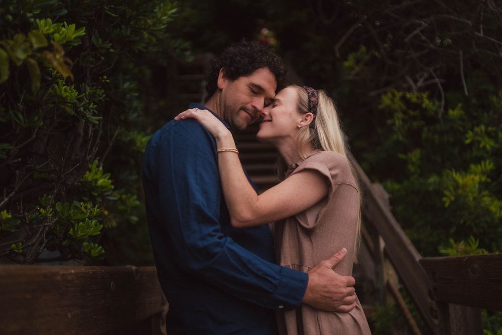A couple embraces in a romantic moment on a wooden bridge surrounded by lush greenery.