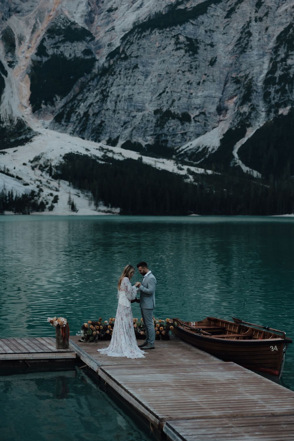 A romantic couple stands on a wooden dock by a wooden boat on turquoise lake waters surrounded by snowy mountains.