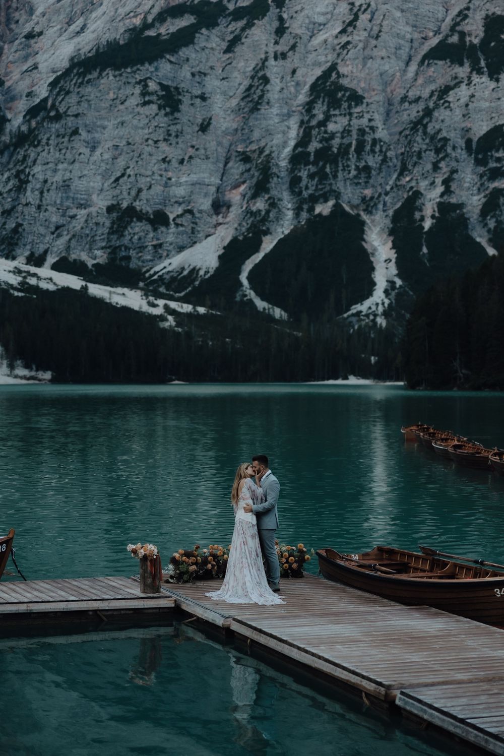 A couple shares a romantic moment on a wooden dock overlooking turquoise Lago di Braies in the Dolomites with wooden boats nearby.