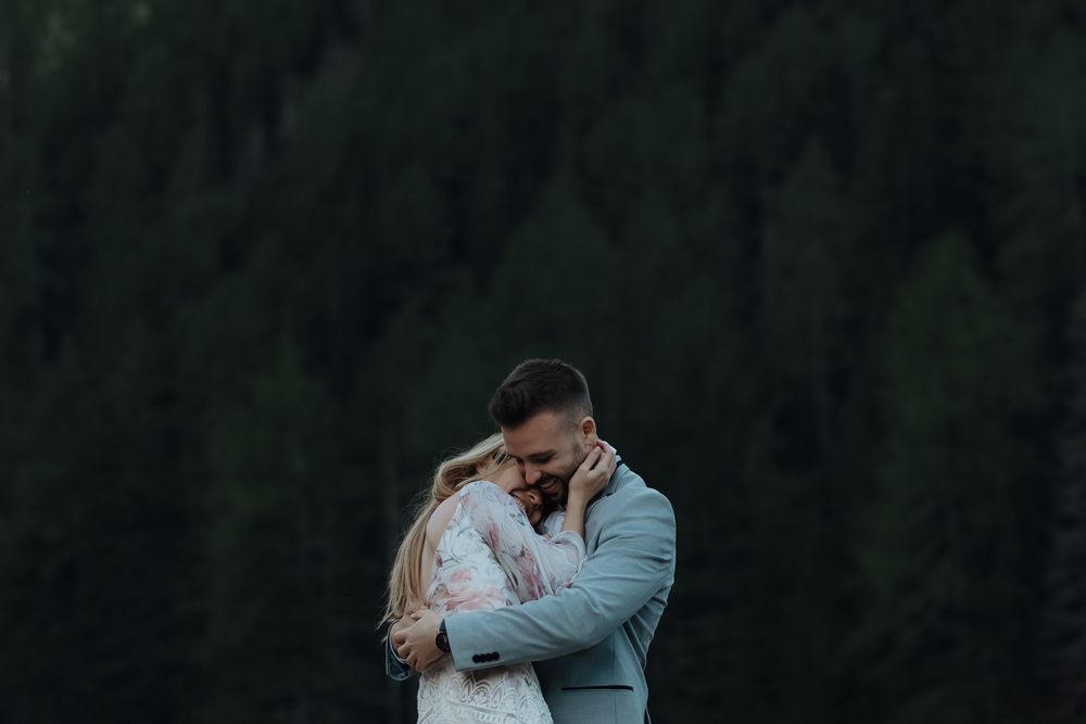 Couple embraces intimately against dark forest backdrop in soft evening light.