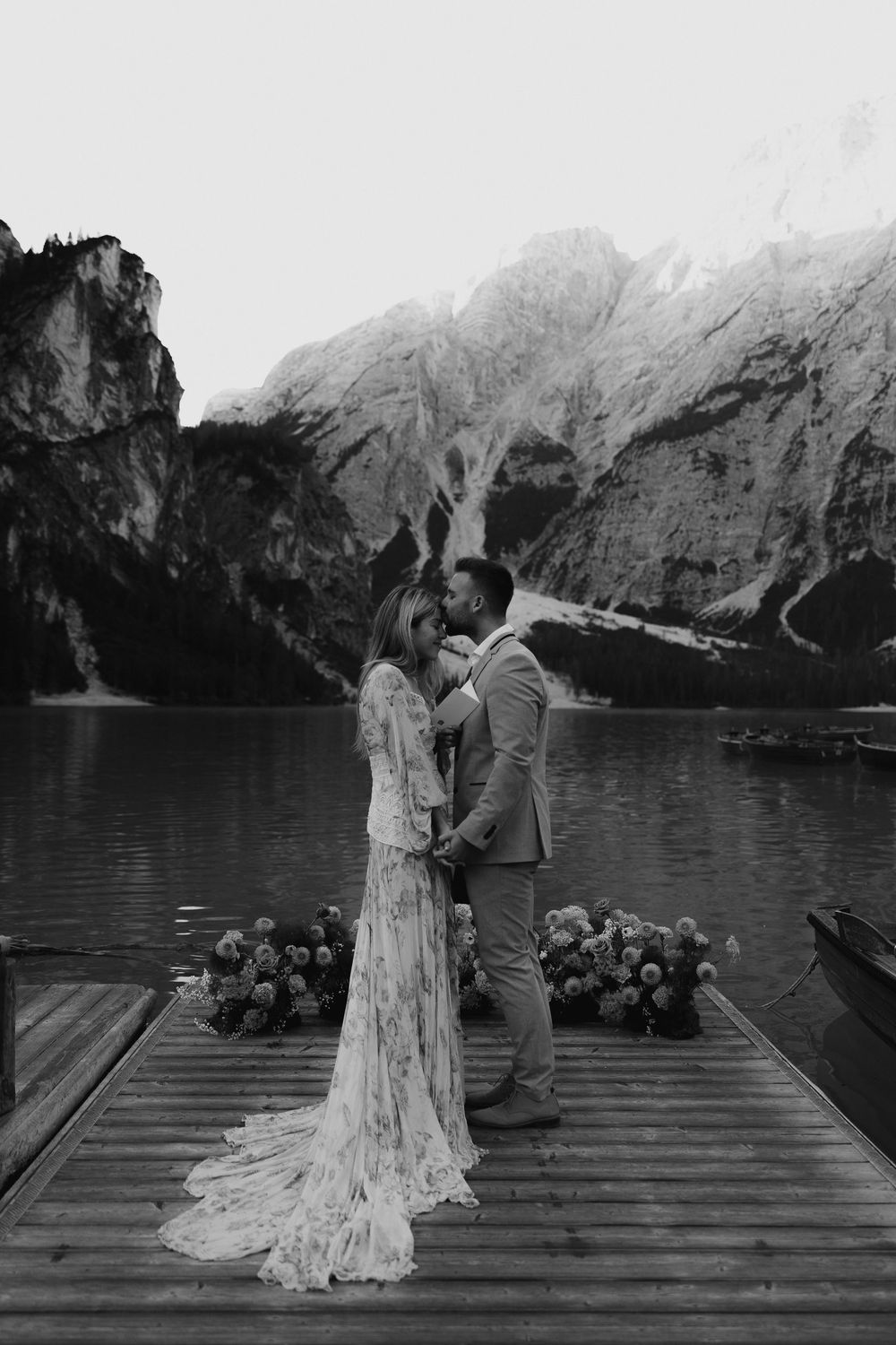 Two figures stand intimately on a wooden dock overlooking a fjord surrounded by dramatic mountain peaks in black and white.