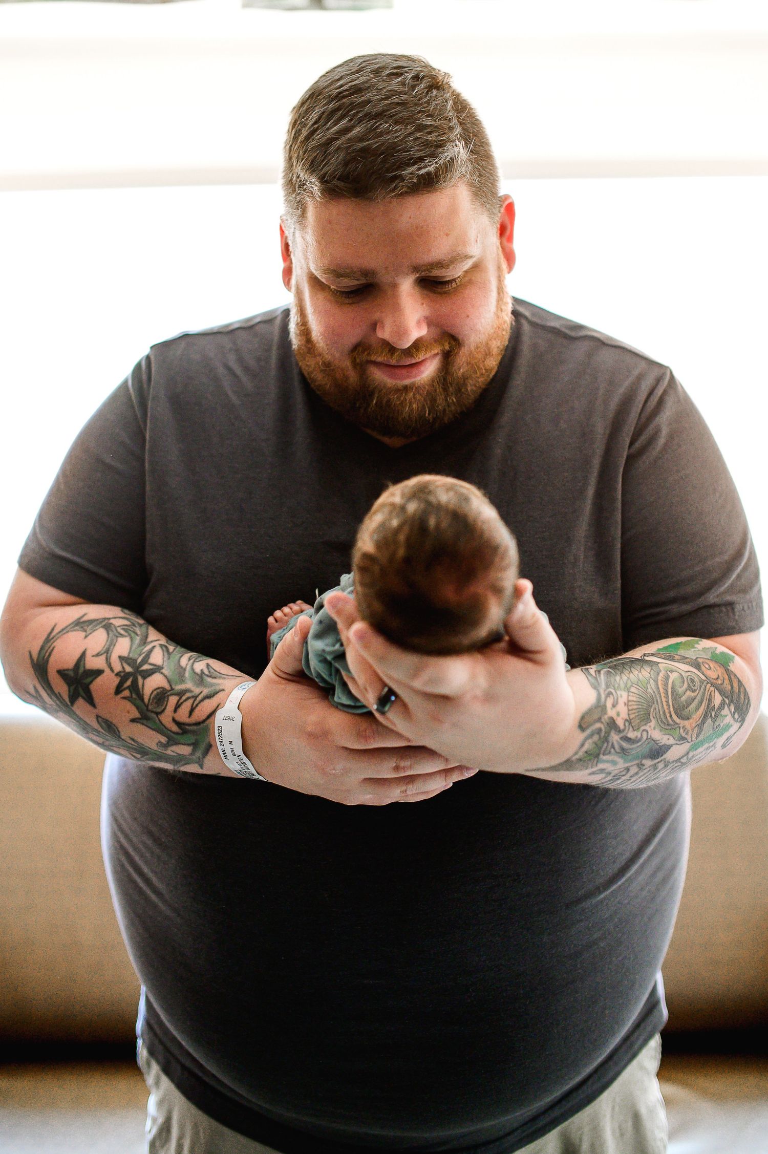 A bearded father in a dark shirt with tattooed arms tenderly holds his infant baby against a light background.