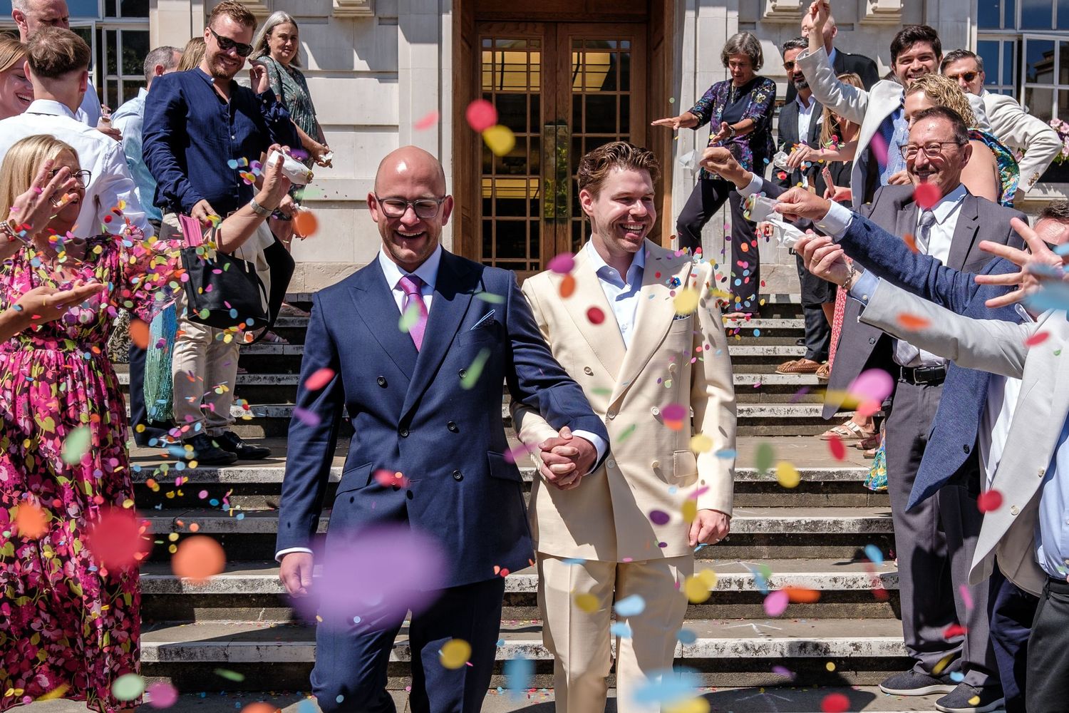 Smiling wedding couple walks down stone steps while guests throw flower petals in celebration.