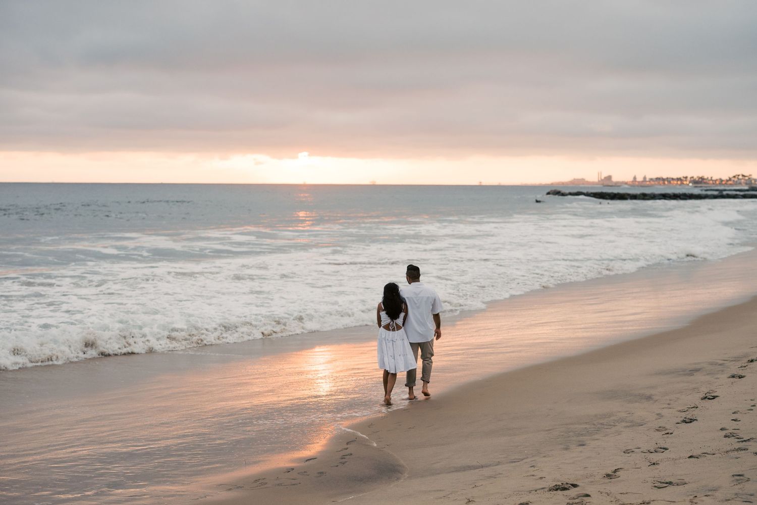 Two people walking hand in hand on a sandy beach at sunset with waves crashing and pink sky.