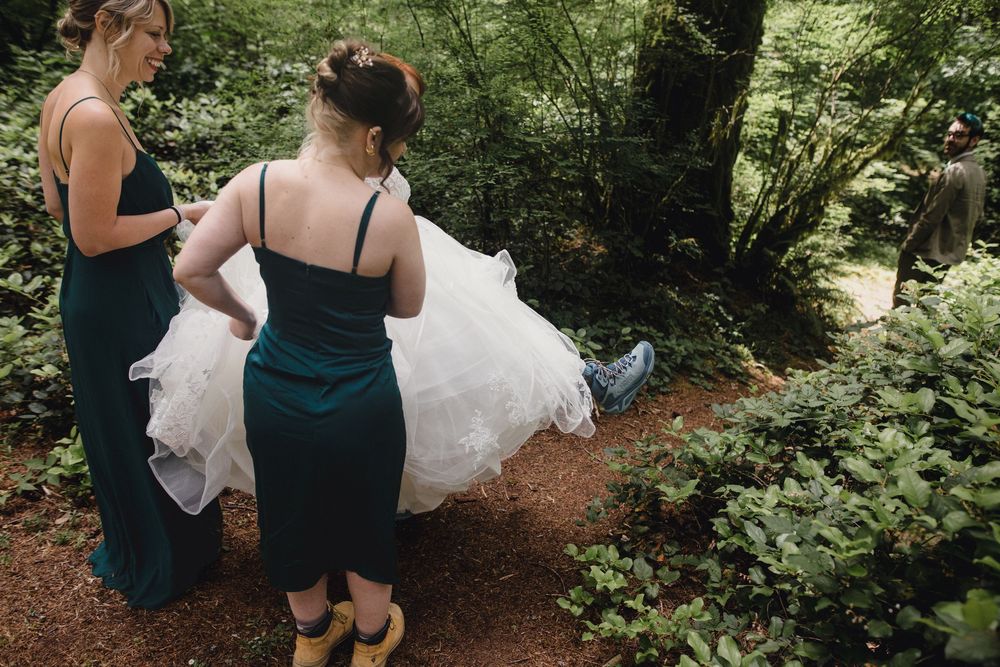 Bridesmaids in dark dresses help arrange a white wedding dress train on a forest path surrounded by greenery.