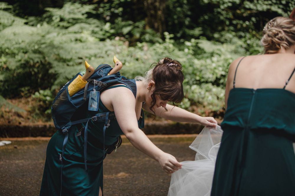 Someone helps adjust a long flowing dress outside in a woodsy setting while wearing a dark green formal gown.