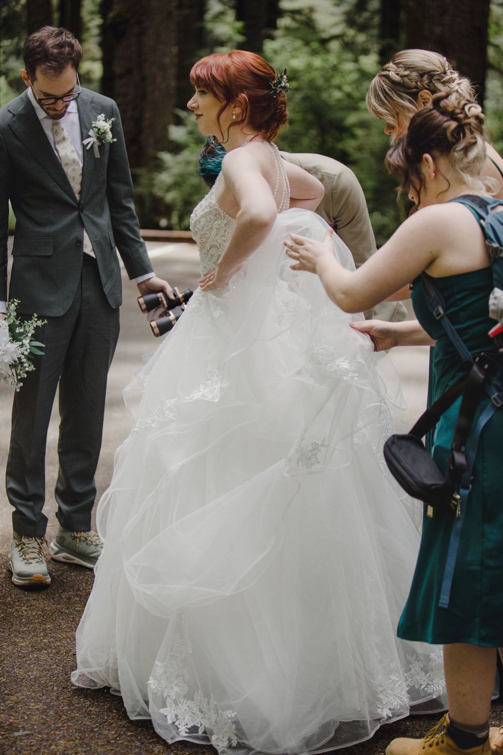 Bride in flowing white gown with bridesmaids in teal dresses gather during outdoor forest wedding ceremony.