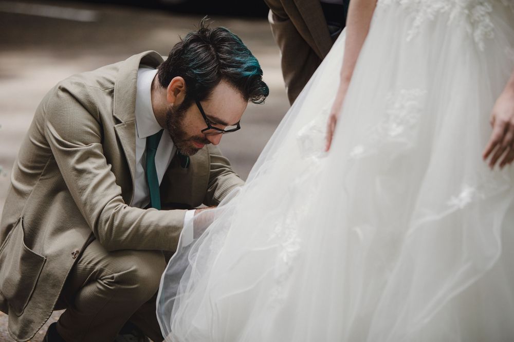 A gentleman in a beige suit and glasses adjusts the hem of a flowing white wedding gown.