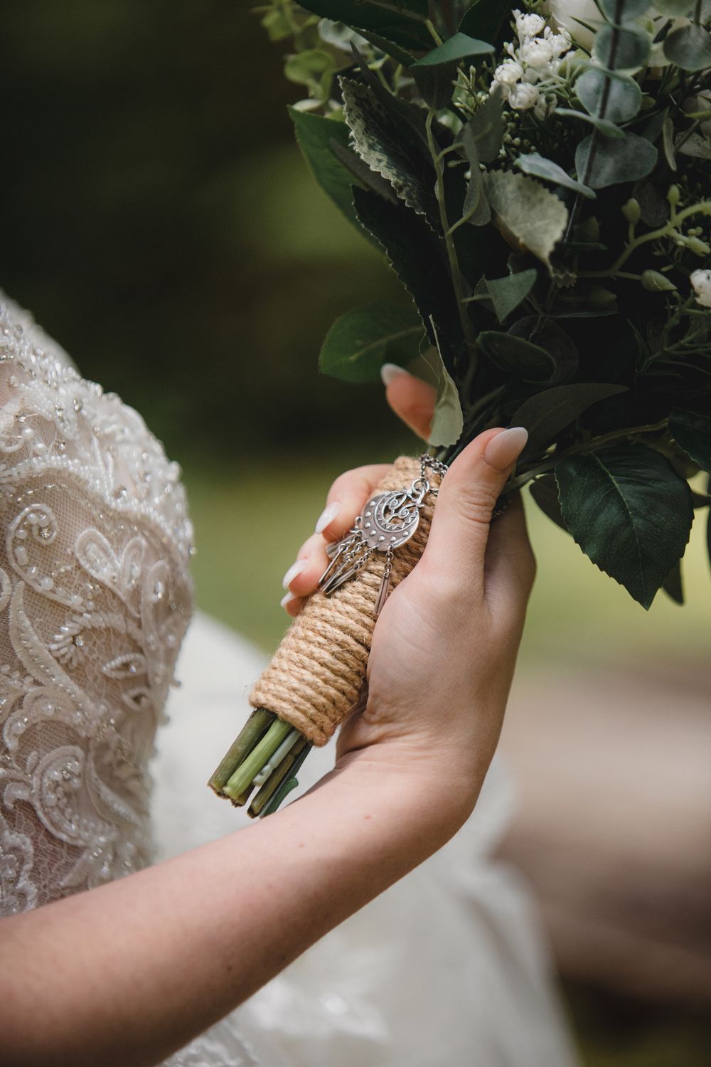Close-up view of hands holding a rustic wedding bouquet wrapped with twine, showcasing lace dress details and manicured nails.