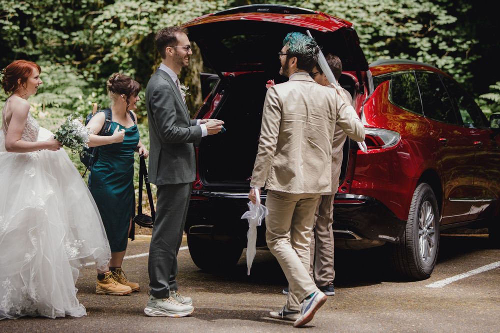 Wedding party standing near a red car in a wooded area during an outdoor wedding celebration.