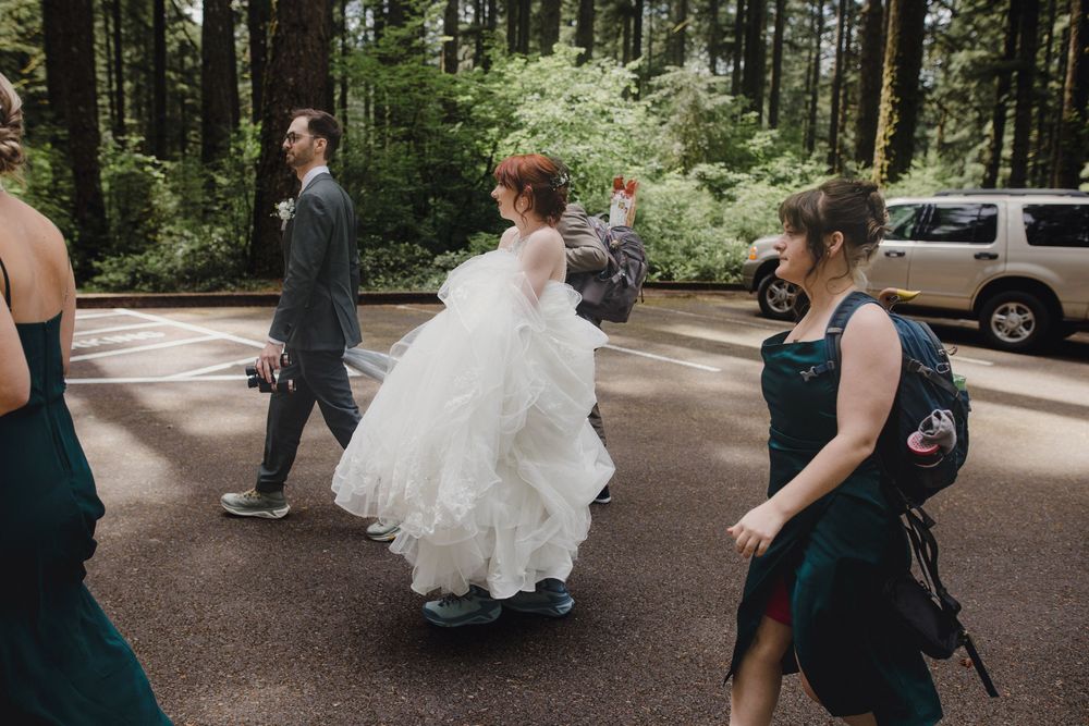 A group walks through a forest parking area with a bride in a flowing white dress.