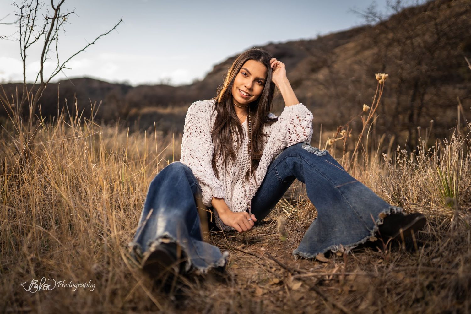 A person in a white lace top and jeans sits casually in tall autumn grass during golden hour.