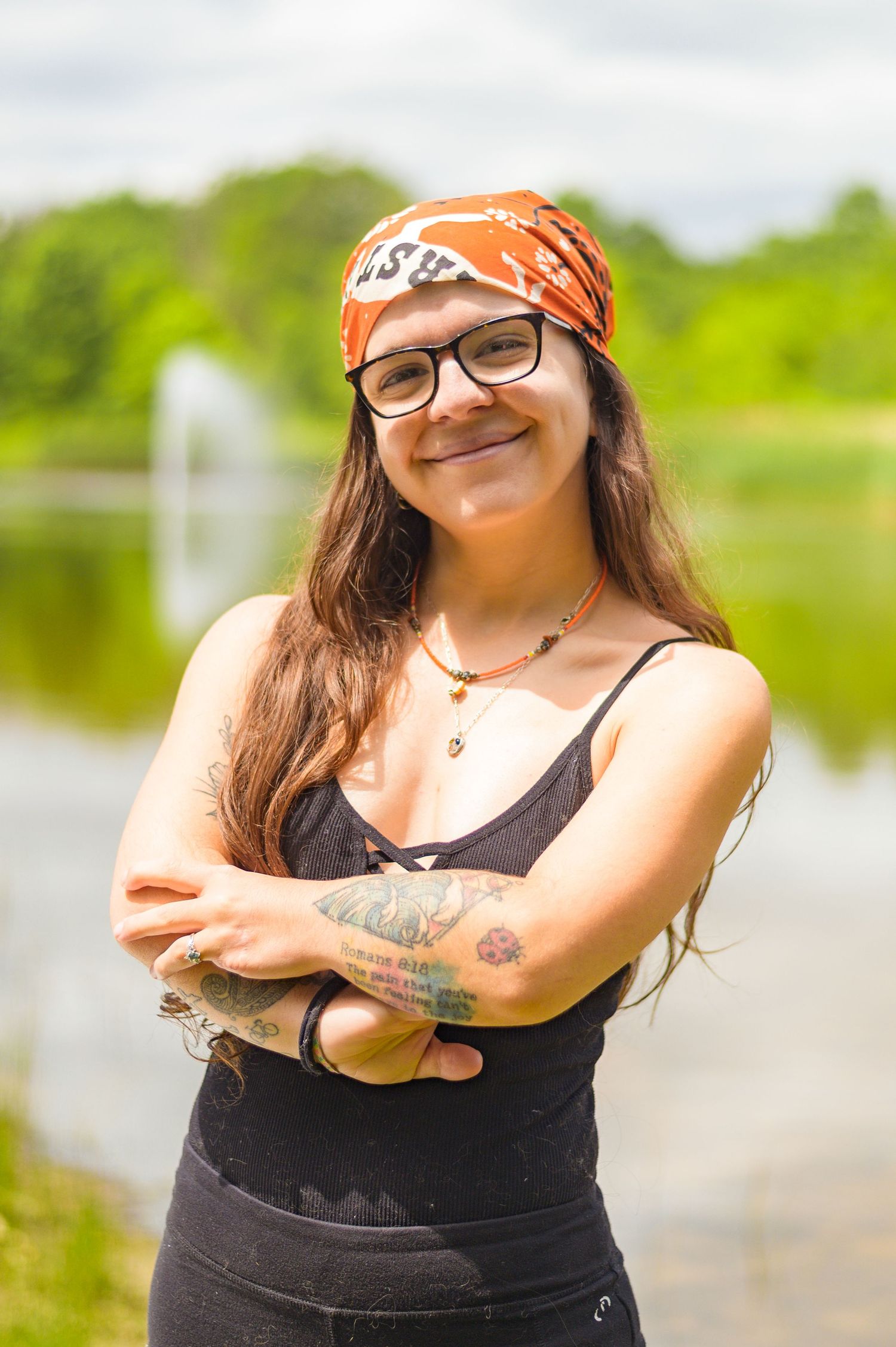 Person in bandana and glasses smiles warmly while standing by a lake.