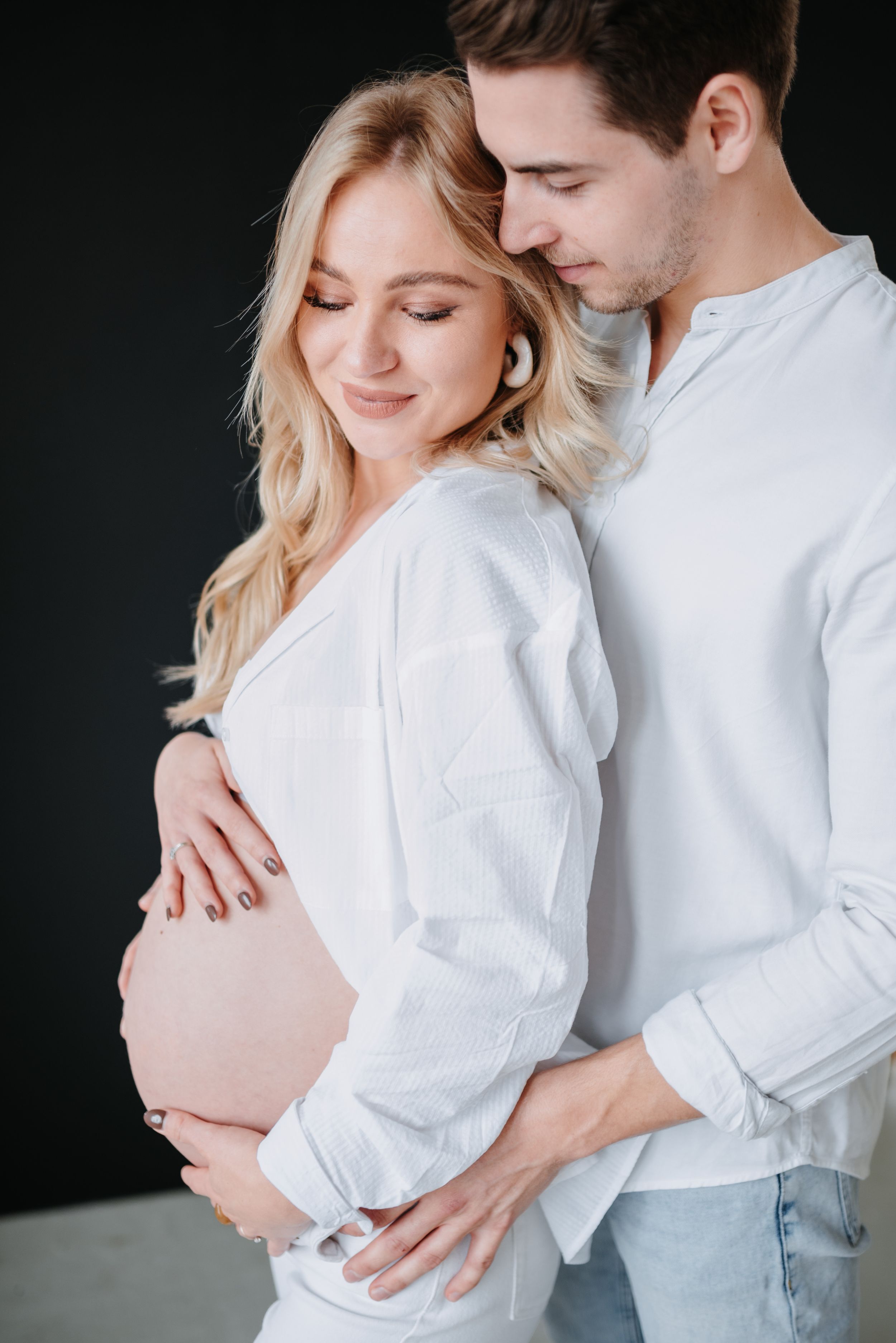 Tender maternity moment features couple in white shirts embracing against dark background as they await their baby.