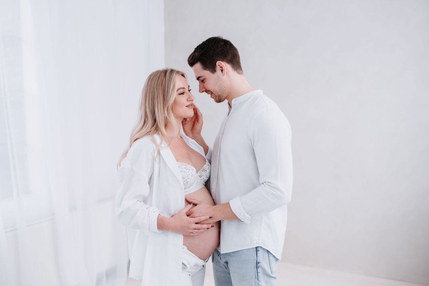 Romantic couple wearing white embraces and kisses tenderly during intimate maternity photoshoot.