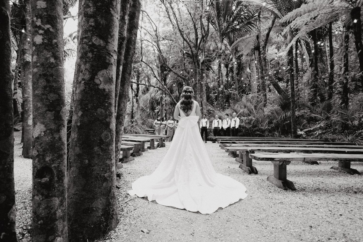 A black and white photo shows a wedding gown from behind as the groom waits captured by Apertured Media Auckland Wedding Photographer.