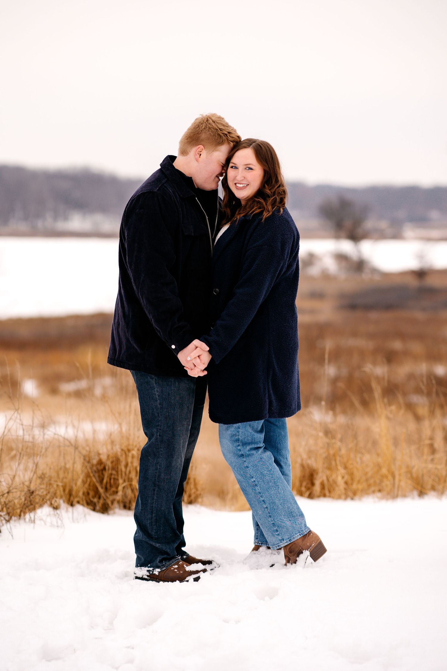 A romantic winter portrait of a couple embracing in a snowy field with a lake in the background.
