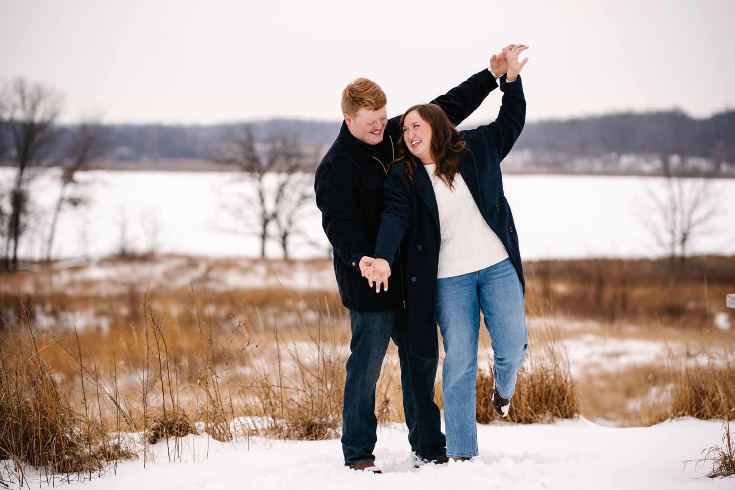 A couple dancing together in a snow-covered field with dried grass and a lake view.