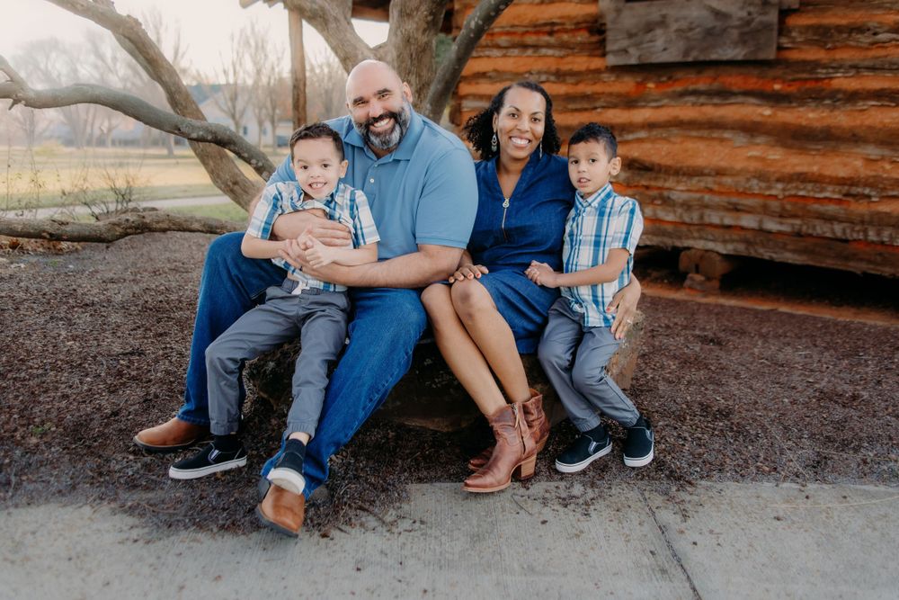 A family sits together outdoors against a rustic wooden wall on a casual afternoon photo shoot.