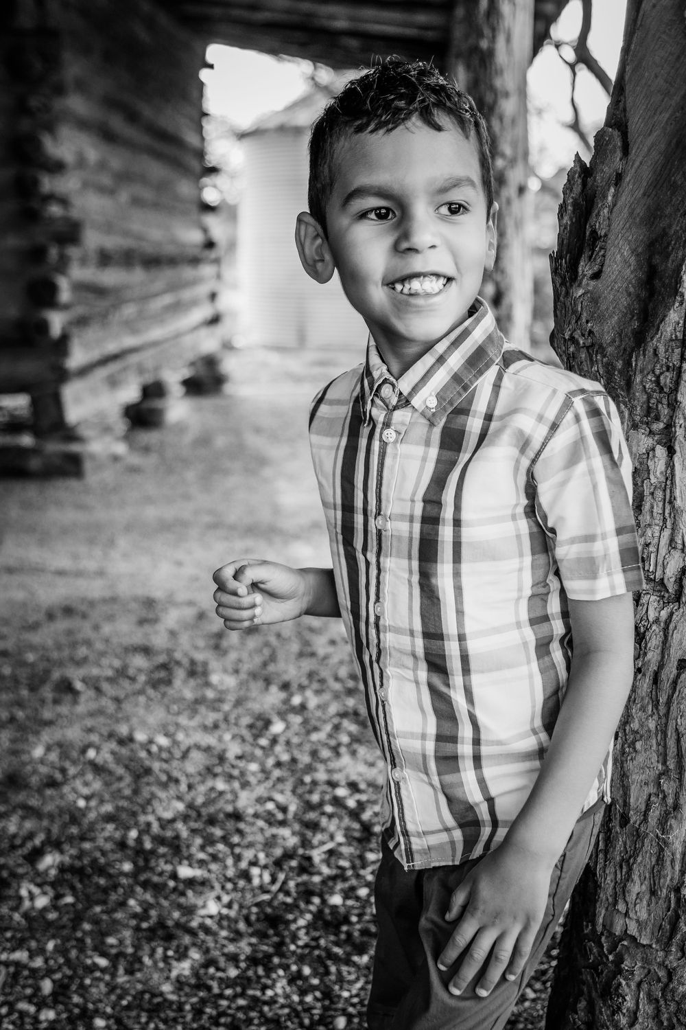 Black and white portrait of young boy in plaid shirt leaning against tree.