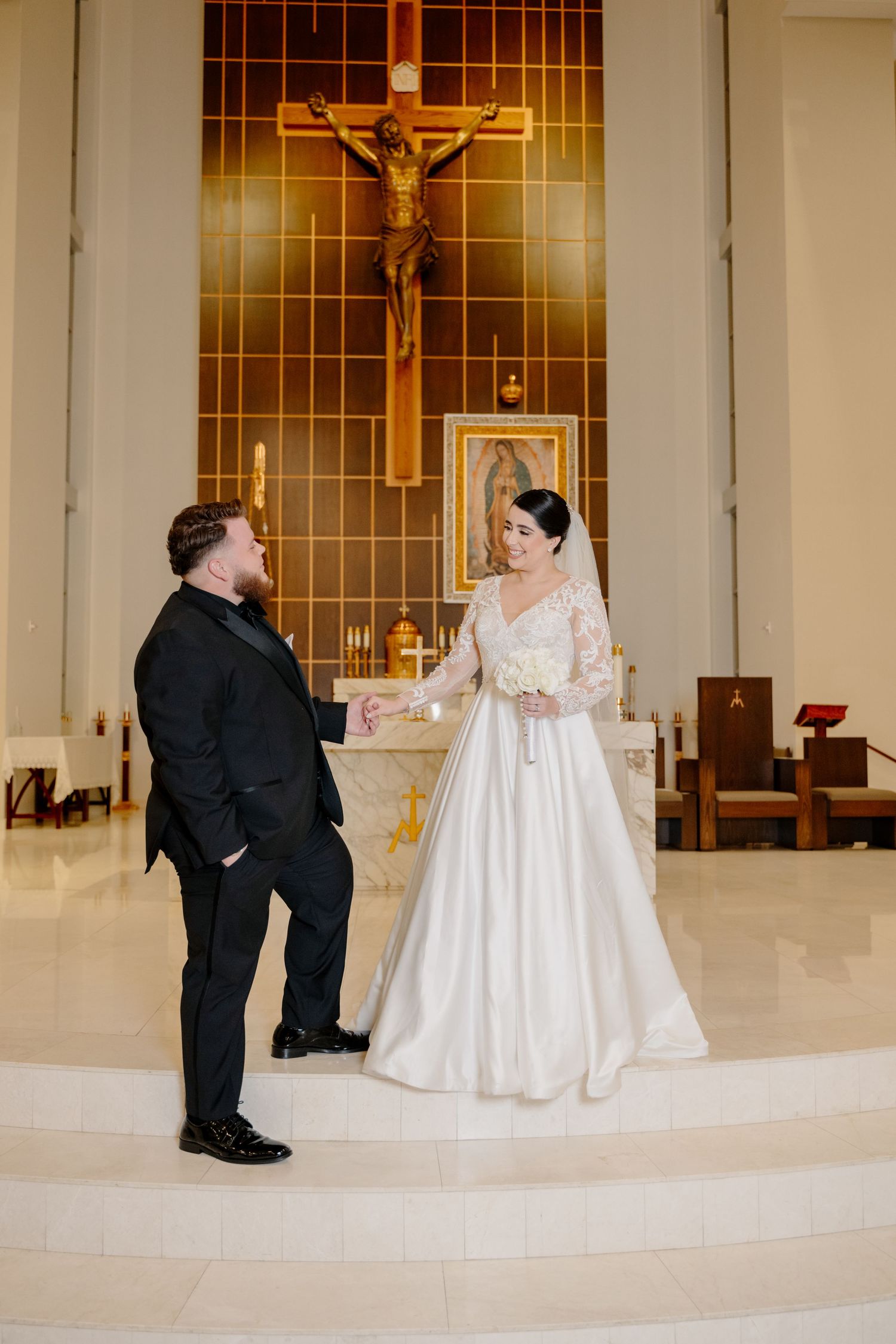 A couple exchanges vows during their wedding ceremony in a church with a large crucifix and warm lighting in the background.