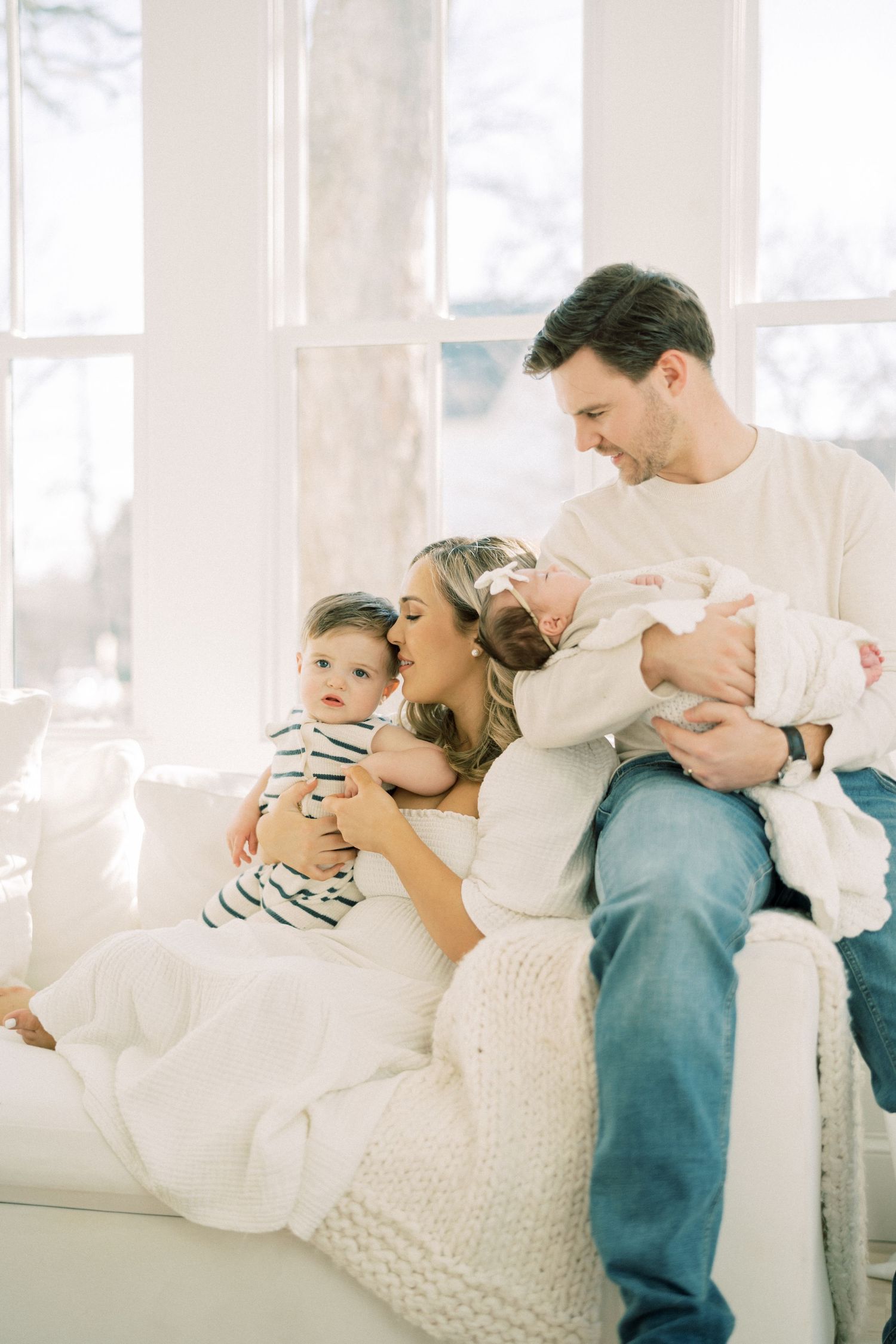 A family relaxes together on a bright white couch by large windows in a tender moment at home.