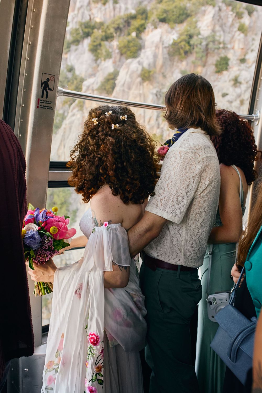 Group stands together looking out a window at mountain cliffs while holding colorful floral bouquets.
