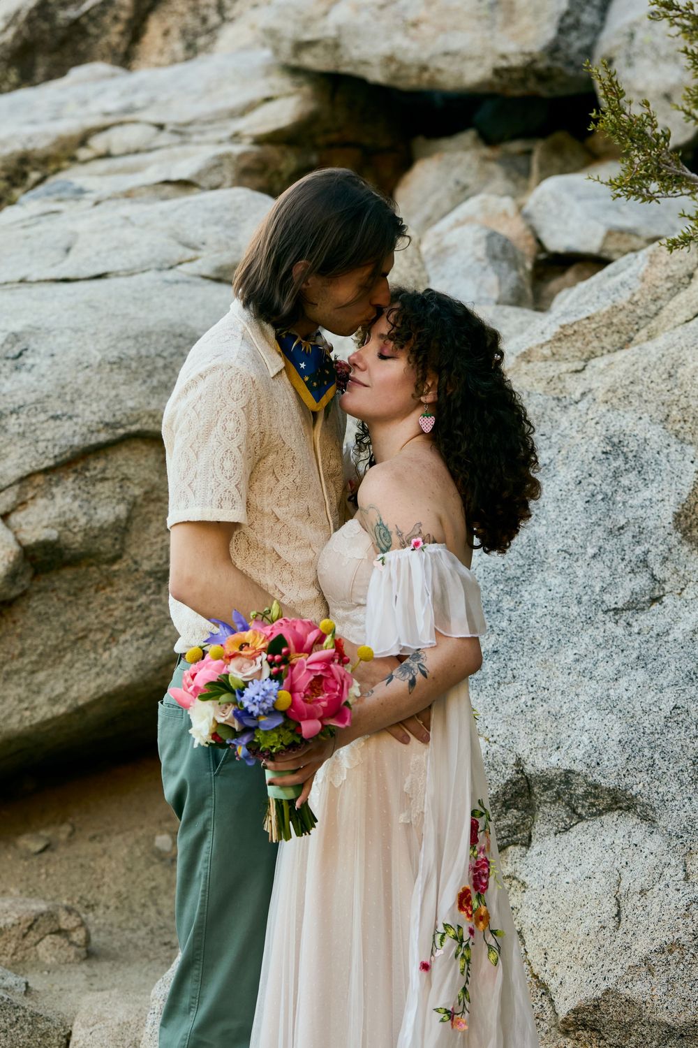 A couple in romantic embrace holds colorful bouquet against rocky cliff backdrop during outdoor photoshoot.