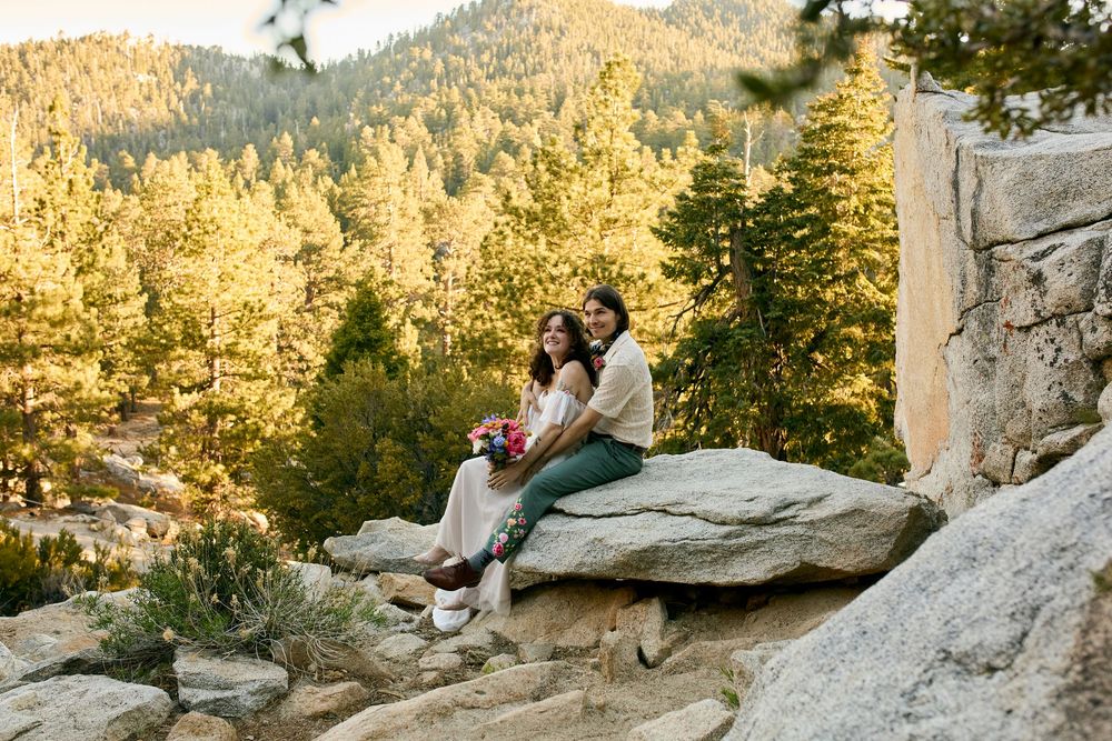 Couple sitting together on rocky outcrop surrounded by golden sunlit evergreen forest in the mountains.