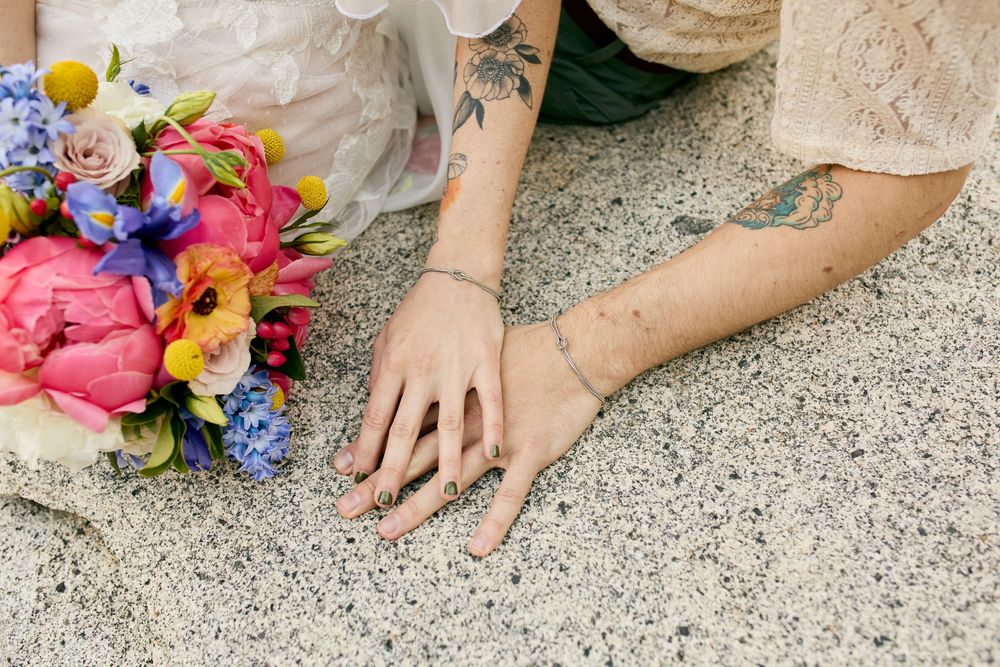 Hands with delicate tattoos rest on concrete beside vibrant coral and blue floral wedding bouquet.