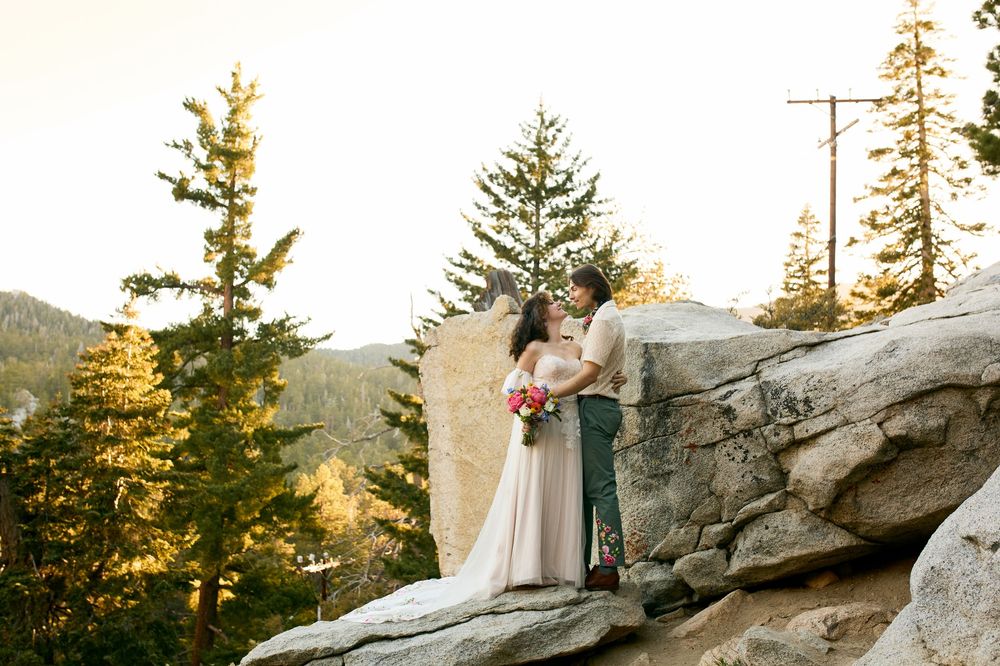 Romantic couple stands on mountain cliff edge surrounded by evergreen trees in golden sunset light.