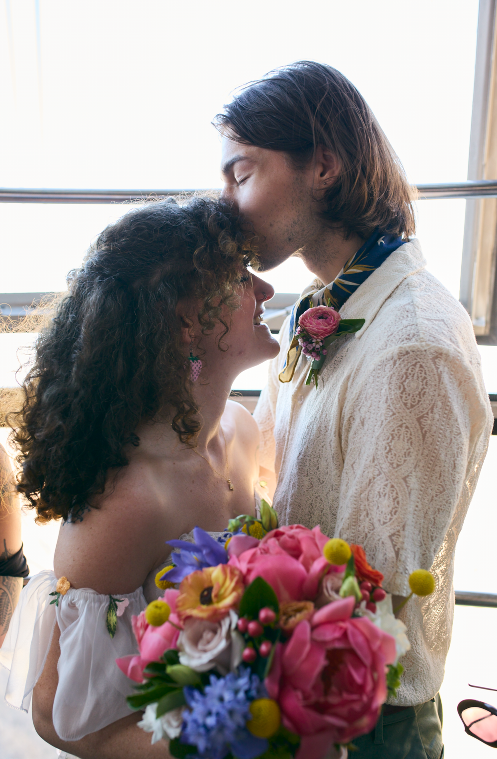 Romantic wedding couple shares tender moment by window with colorful floral bouquet of pink roses and purple blooms.